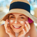 Donna sorridente con cappello e crema solare sul volto